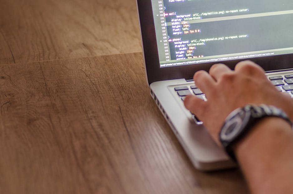 The image shows a person's hand with a wristwatch, typing on a laptop keyboard placed on a wooden surface. The laptop screen displays lines of code or text, indicating digital work or programming acti